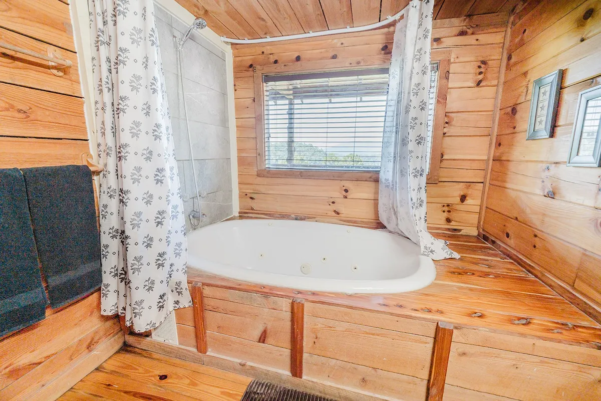 A luxurious jacuzzi tub sits beside a large window framed by wood paneling and decorative curtains, with soft daylight pouring in and the tranquil outdoors visible. The inviting setup promises a rejuvenating soak after a day of hiking or exploring.