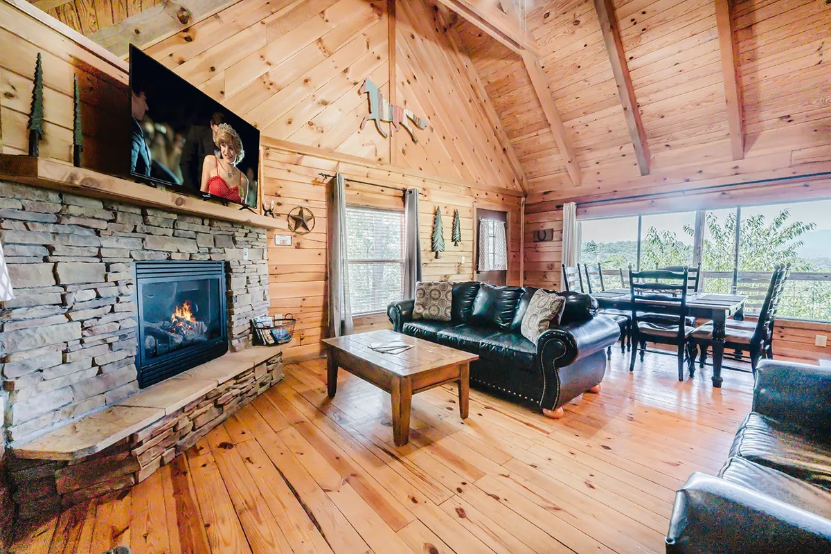 A comfortable living area with a plush sofa near a crackling stone fireplace, adjacent to a dining space and a wall-mounted TV. The space glows with natural light, suggesting a seamless flow between lounging, dining, and socializing.