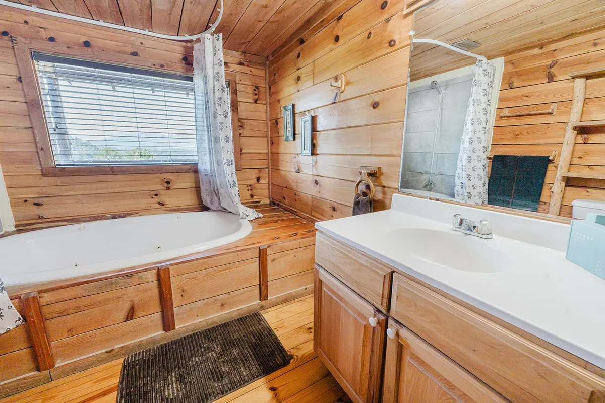 A spacious bathroom with a deep bathtub, rustic wooden wall panels, a wide sink, and a window providing natural light and views. The atmosphere is tranquil and spa-like, perfect for relaxing routines.