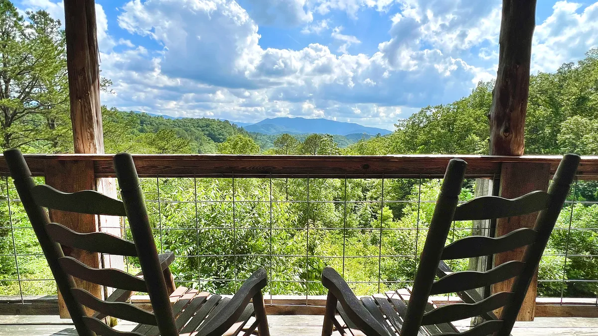 The image features a pair of classic wooden rocking chairs set on a cabin porch, overlooking sweeping mountain views under a peaceful, cloudy sky. The scene exudes tranquility and invites guests to relax, soak in the natural beauty, and experience quiet moments surrounded by the Smoky Mountains.