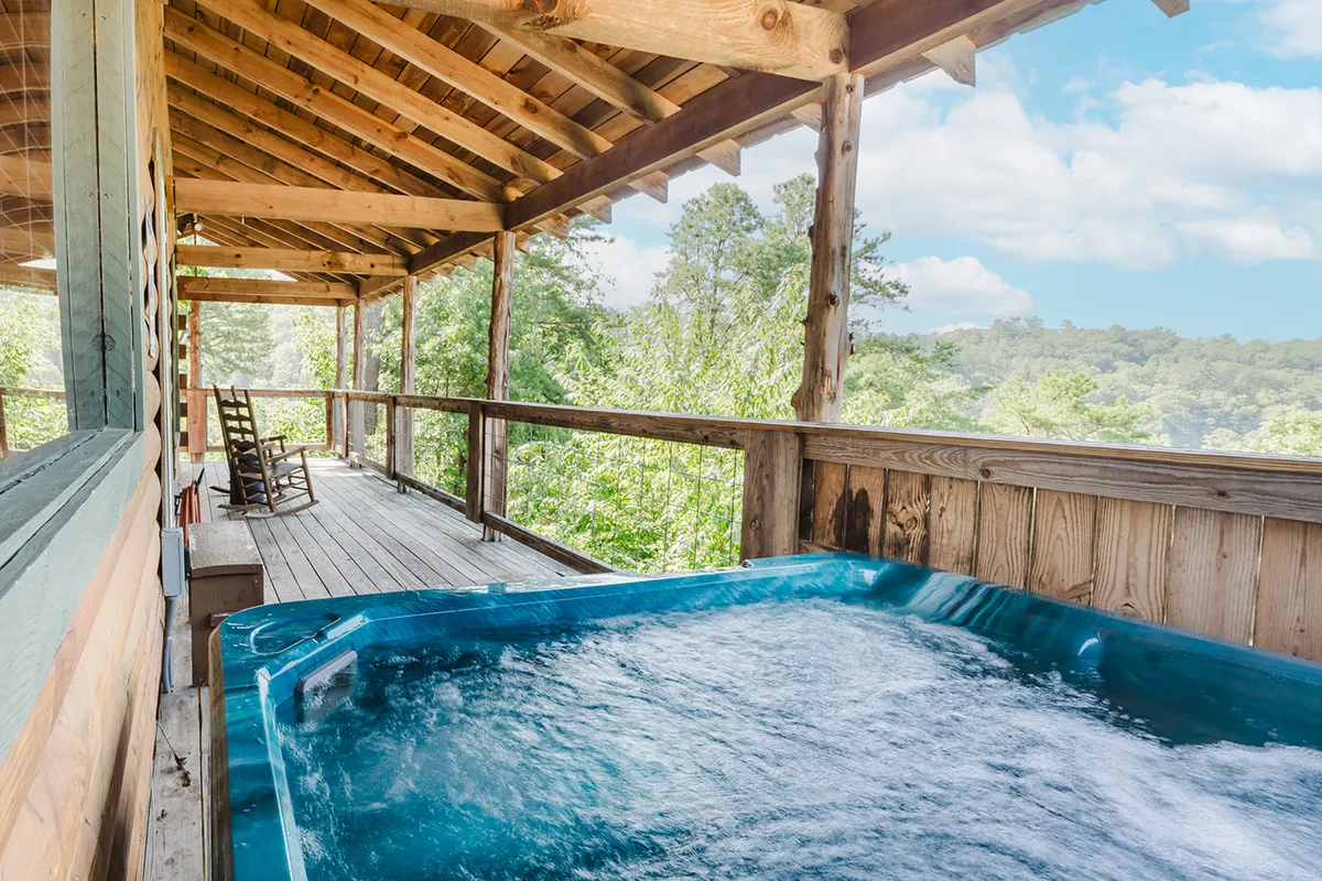 A spacious wooden deck features a bubbling hot tub and rustic rocking chairs, set against a backdrop of lush greenery. The scene suggests relaxing soaks at sunset, morning coffee outdoors, and stargazing after adventure-filled days.