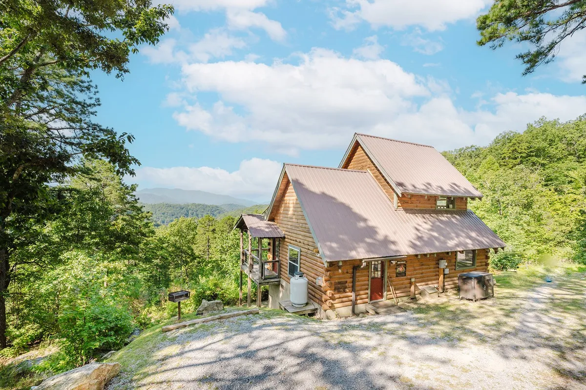 A classic log cabin sits amidst lush trees with the Smoky Mountains rising in the background. The cabin's rustic wood exterior and surrounding greenery set a welcoming scene for outdoor adventure. The wide vistas and natural setting evoke the proximity to national park adventures.