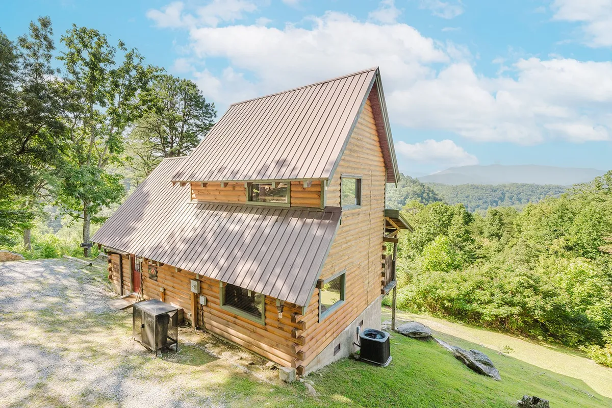The log cabin is framed by tall trees, with a sweeping view of the mountains behind. The porch and outdoor area invite thoughts of relaxing after a day of Smoky Mountain adventures. The setting highlights the cabin’s role as a peaceful retreat after energetic explorations.