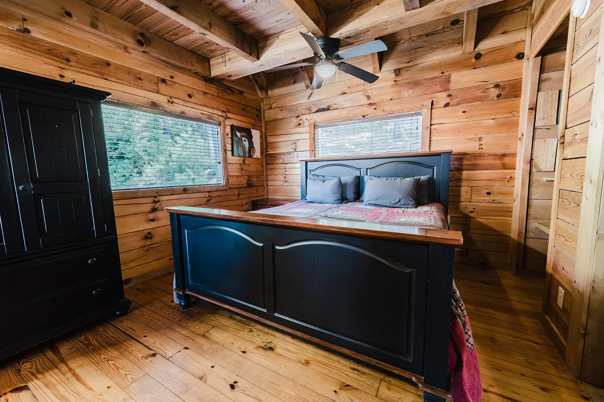 A sunlit bedroom features a wooden bed with plush linens, a black dresser, large windows letting in ample natural light, and a ceiling fan overhead. The space feels inviting and tranquil, perfect for relaxation after a day of adventure.