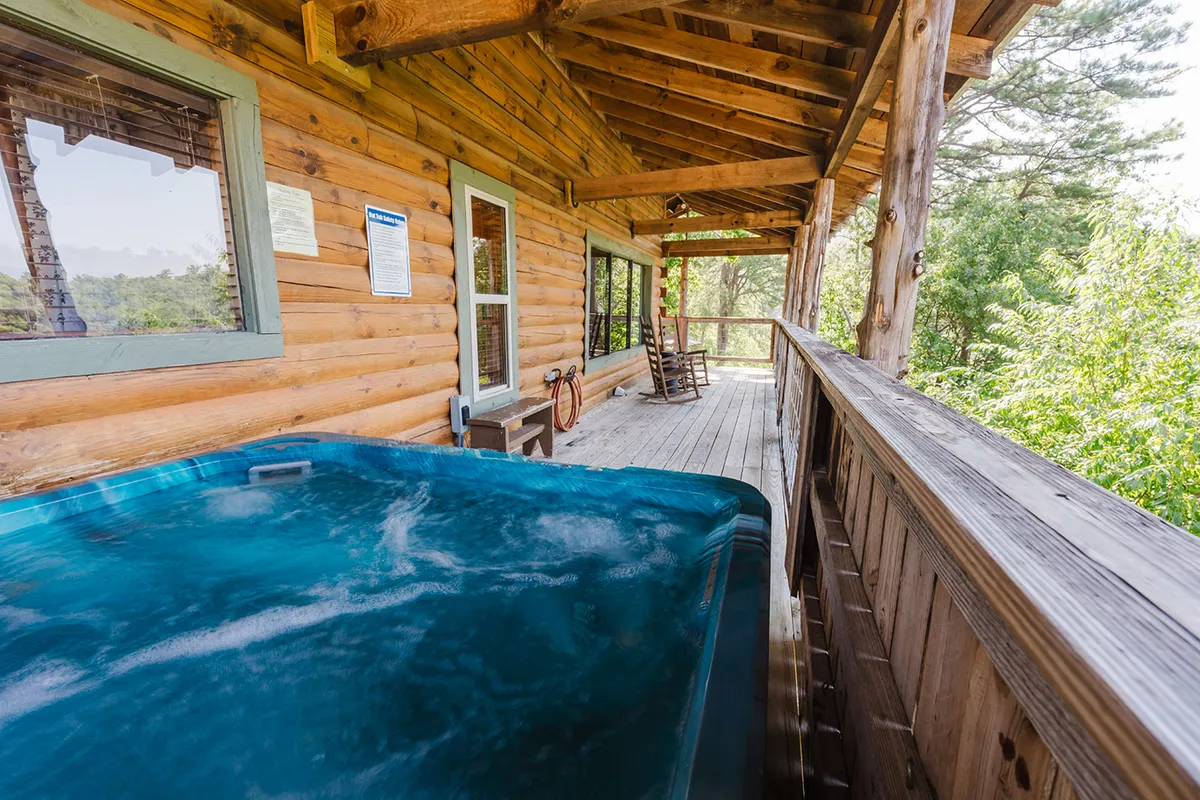 A view of the wooden deck featuring a bubbling hot tub, comfortable rocking chairs, and the rustic logs of the cabin. The scene is tranquil, with sunset light filtering in and panoramic mountain vistas stretching into the distance. Friends can be seen relaxing in the hot tub or unwinding on the deck.