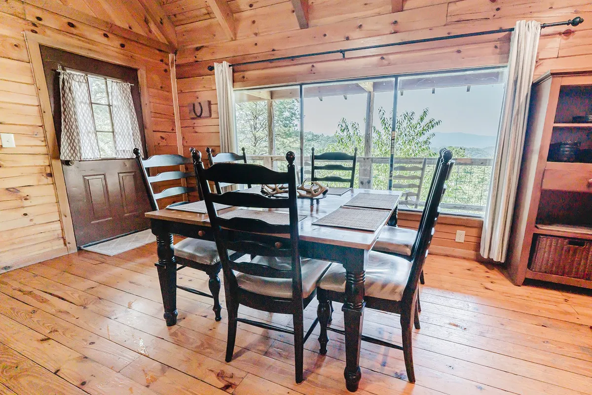 A rustic wooden dining table positioned by a large window that frames scenic mountain views, paired with wooden cabinets and warm lighting. The setting signals a space ideal for family meals, board games, or enjoying coffee while gazing at nature.