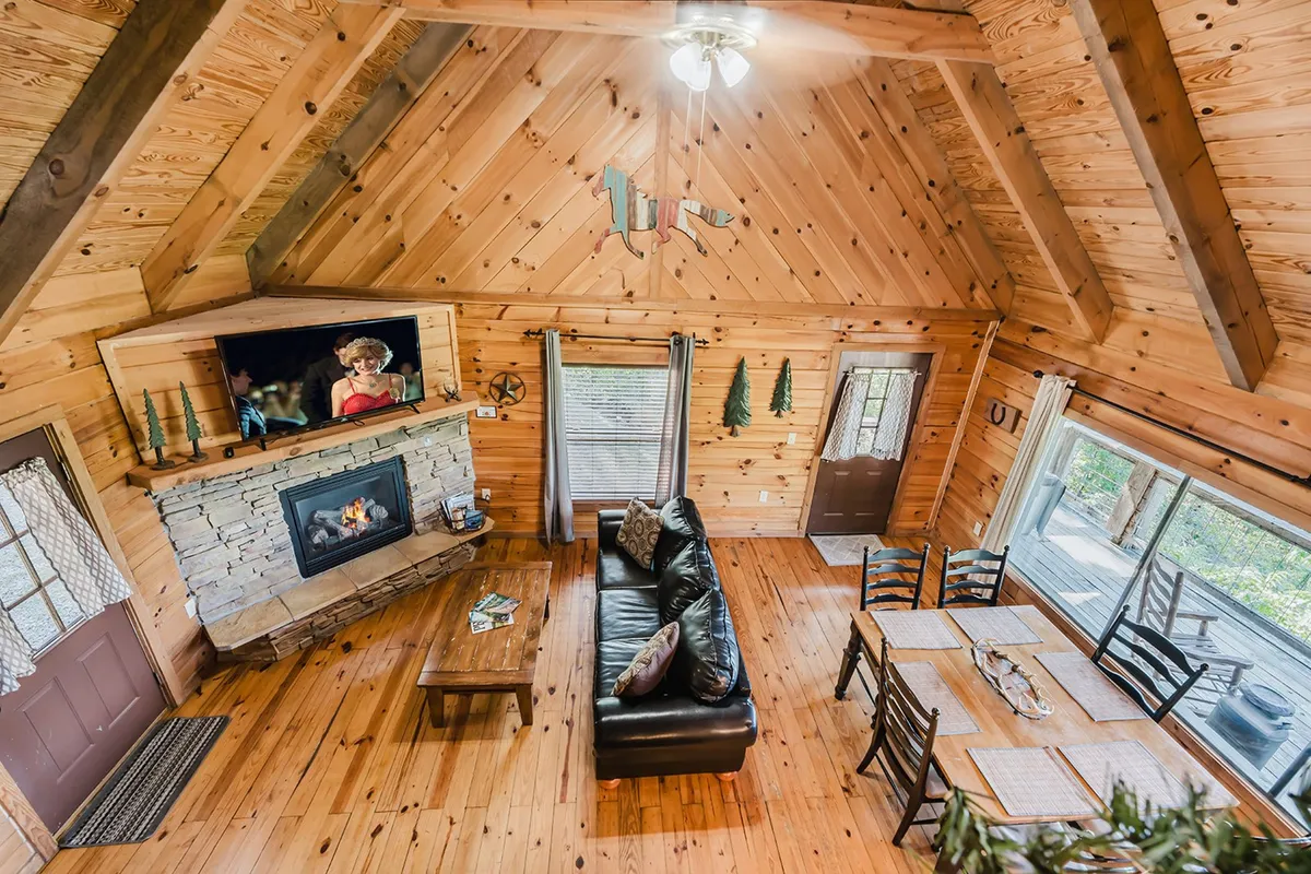 A spacious open-plan living and dining area featuring a stone gas fireplace, large windows offering breathtaking mountain views, a cozy couch with cushions, and a flat-screen TV. The dining table seats six, and sunlight streams in, enhancing the warm and welcoming ambiance.