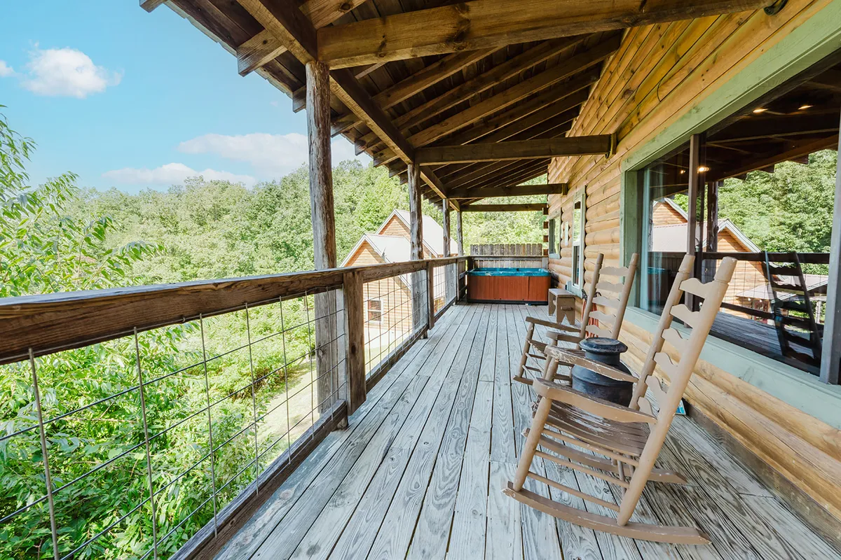 A cozy porch features rocking chairs and a wooden railing, with lush greenery and a hot tub in the background. The setting is peaceful and inviting, perfect for enjoying coffee at sunrise, relaxing after a trail walk, or sharing quiet moments with a pet. The outdoor space feels both private and open to the scenic landscape.