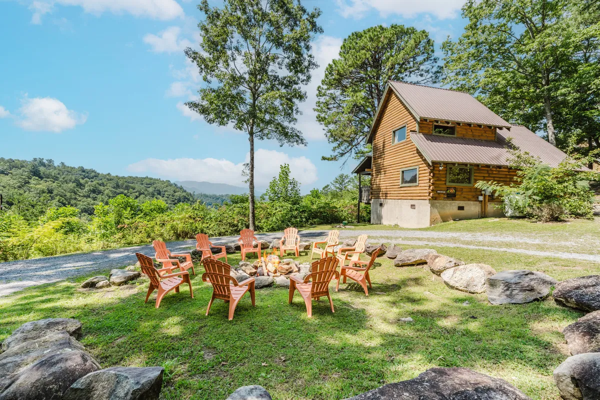 An inviting outdoor fire pit surrounded by classic Adirondack chairs sits just steps from a charming wooden house, all nestled within lush greenery. The setting is ideal for campfire gatherings, storytelling, and making s'mores under the stars with friends, family, and pets.